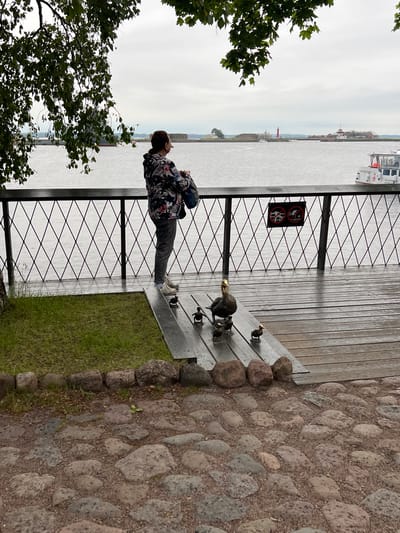 The image depicts a woman standing on a wooden deck overlooking a body of water. She is facing away from the camera, looking towards a distant shoreline with buildings and a lighthouse. She is wearing a patterned jacket, grey pants, and white sneakers, and is holding a bag. In front of her, on the deck, are several bronze duck sculptures, one adult and several ducklings. The deck is surrounded by a black metal fence with a diamond pattern. To the left of the deck is a grassy area and a cobbled path. The weather appears overcast. The location is identified as Kronstadt, Russia, likely near the Kronstadt Sea Cathedral or Forts, given the presence of a lighthouse and fortified structures across the water.