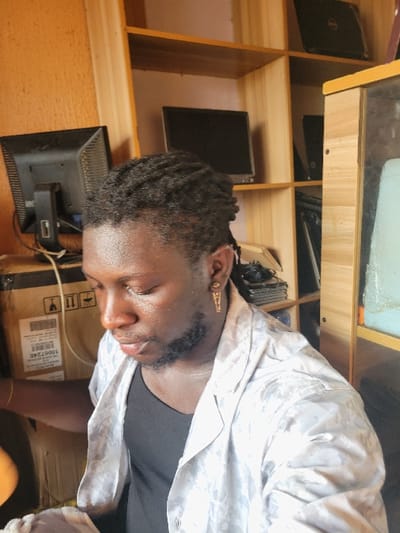The image displays a male individual in an indoor setting within Makurdi, Nigeria, focusing downwards. The subject has dark skin, short dreadlocks, and a trimmed beard. He wears a black undershirt beneath an unbuttoned, light-colored collared shirt with a subtle pattern. A gold-colored earring, possibly an ankh or similar symbol, is present in his right ear, and a gold-toned bracelet adorns his left wrist.
The background consists of a light-brown wooden shelving unit. The top shelf holds two black laptops, one partially visible. The middle shelf displays two black desktop monitors. The bottom shelf contains stacked black electronic components. To the subject's left, a large cardboard shipping box is visible, featuring labels including "10007246" and handling symbols, with a black cable running alongside it. Another black desktop monitor is positioned behind this box. To the subject's right, a light-colored wooden cabinet with a reflective panel showing a faint blue-green tint is present. The wall behind the shelving unit is a solid peach color.
