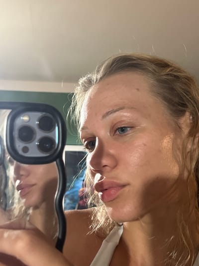 This media file is a close-up, upside-down selfie taken by a young woman with blonde hair. She is looking towards the camera, with her eye visible and a hint of a smile on her lips. Her skin appears dewy, possibly indicating she has applied skincare or makeup. She is wearing a white top. The background is out of focus, but hints of a room with green walls are visible. A phone with a distinctive camera bump is held by the subject, reflecting her face and upper body. The reflection shows her blonde hair and the white top. The lighting suggests it is daytime, possibly indoors. The overall mood is casual and intimate. No visible text is present in the image. The location context is Mühldorf a. Inn, Germany.