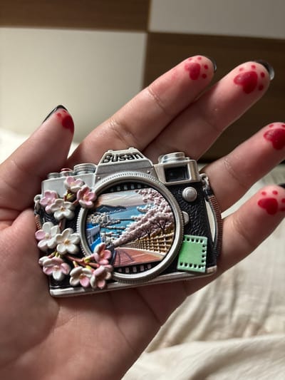 The image shows a close-up of a person's hand holding a camera-shaped magnet. The magnet features a scene of a city with cherry blossoms and water. The person's fingers have red paw print designs. The background appears to be a bed or a similar soft surface, with a hint of furniture in the upper portion of the image. There is no visible indication of the city of Shenyang, China in the image.