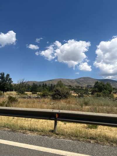 The image depicts a roadside view of a rural landscape with rolling hills and scattered trees under a bright blue sky with fluffy white clouds. In the foreground, a dark grey metal guardrail runs horizontally across the frame, separating the asphalt road with a white stripe on the left from a dry, grassy embankment on the right. The embankment is sparsely populated with vegetation, including tall, dry grass and some scattered green bushes.
Further back, the landscape opens up to reveal a field of dry, golden grass, dotted with more green shrubs and trees. A low stone wall or fence is visible in the mid-ground, running along the edge of the field. Behind this, a variety of trees, including evergreen conifers and deciduous trees with sparse foliage, are scattered across the terrain. The background is dominated by a range of gently sloping, arid-looking hills, appearing rocky and sparsely vegetated. The overall impression is of a dry, sunny day in a somewhat undeveloped, hilly region.
The image is consistent with a view from a road passing through the outskirts of El Espinar, Spain. El Espinar is known for its varied landscape, which includes mountainous areas and dry, grassy plains, particularly during the warmer months. The presence of a guardrail suggests the image was taken from a road, and the natural, somewhat rugged terrain is characteristic of the region. There are no visible people or specific landmarks that pinpoint an exact location within El Espinar, but the general environmental features align with the typical scenery found in and around the municipality.