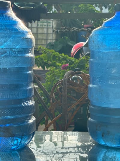 The scene is set outdoors in Ko H'Neh, Vietnam, observed through a reflective surface from a slightly elevated perspective. In the immediate foreground, two large, clear blue plastic water bottles, exhibiting condensation, flank the central view, resting on a wet or reflective surface that shows distorted reflections of the background. Between these bottles, a densely packed outdoor environment is visible.
A prominent mass of vibrant green foliage occupies the midground, featuring a single fuchsia-colored flower towards its center. Partially obscured by the foliage, a coiled orange hose is visible, along with metallic components of what appears to be machinery, possibly a water pump or pressure washer. Behind these elements, a red motorcycle helmet rests on a dark, indistinct object, likely a motorcycle. Further back and to the left, a large white plastic storage tank is partially visible. Above the foliage and machinery, a horizontal metal bar structure is present, and a twisted, gnarled branch extends into the frame. The overall lighting suggests daylight. No people are directly visible, but a faint, distorted human-like silhouette is reflected on the surface below the left water bottle.