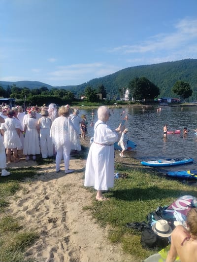 The image depicts a sunny day at the lake in Międzybrodzie Bialskie, Poland. A group of people, predominantly women dressed in white attire, are gathered on the sandy shore. Some are wearing floral headpieces. In the background, the lake is populated with swimmers, some of whom are using inflatable rings. Paddleboards are visible near the shore. The landscape is characterized by green hills and trees under a clear blue sky. Personal belongings, including bags and hats, are scattered on the grass near the shore. One woman in the foreground is taking a photo with her phone, while another person in white is holding a fan.