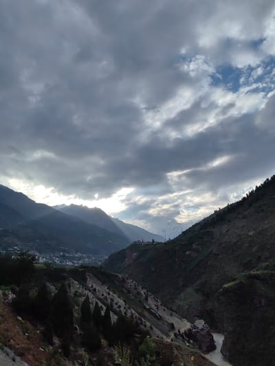 This is a landscape photograph taken in Kardang, India, showing a valley carved by a river. The scene is dominated by imposing mountains that rise on either side of the valley. The sky is filled with dramatic clouds, with shafts of sunlight breaking through, illuminating parts of the mountainside. In the distance, a snow-capped peak is visible. On the slopes of the mountains, scattered settlements with buildings are discernible. A winding river flows through the valley floor. The vegetation on the mountainsides includes coniferous trees and sparse undergrowth. The overall impression is one of rugged natural beauty and a remote, mountainous environment.