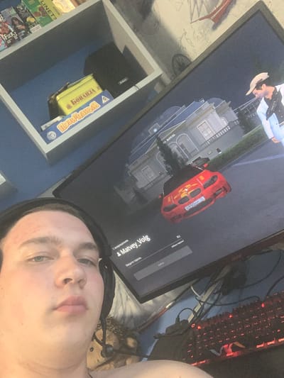 The image shows a young man, likely a teenager or young adult, taking a selfie with a computer monitor in the background. The man is positioned in the foreground, occupying the left side of the frame, and is wearing headphones. The monitor displays a video game interface, featuring a red sports car and a building with the text "ROYAL" visible. On the left side of the image, a shelf unit contains books and board games, including one with Cyrillic text that appears to be "Бонanza" (Bonanza). The overall environment suggests a personal gaming setup within a home. There is no information present in the image that would allow for the identification of the specific location in Severodvinsk, Russia.
