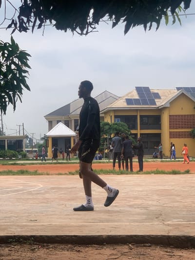 The image depicts an outdoor scene, likely a sports field or court, with a building in the background. Several people are visible, engaged in what appears to be a sports activity or training. The primary subject in the foreground is a young man in athletic attire, a black t-shirt and shorts, with white socks and dark athletic shoes. He is in motion, his body positioned as if mid-stride or jump, with one leg extended and the other bent.
In the background, a yellow building with solar panels on its roof is prominent. A group of other individuals can be seen near the building, some in athletic wear, suggesting a sporting event or practice. The ground is a mix of reddish-brown dirt and a lighter colored surface, possibly a court. Lush green foliage is visible on the left and bottom edges of the image, framing the scene. The sky appears overcast, suggesting a cloudy day. The time of day is likely daytime, given the lighting conditions. There is no discernible text in the image. The overall atmosphere seems to be one of athletic activity and training in a somewhat casual, outdoor setting.