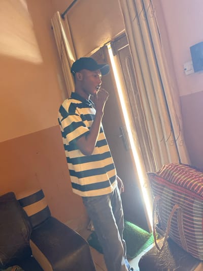 An indoor scene depicts a male individual standing near a brightly lit doorway in a room with light brown walls. The individual, positioned slightly right of center, faces right in profile, wearing a black baseball cap, a navy blue and white horizontal striped short-sleeve polo shirt, and dark trousers. His right hand is raised towards his mouth or chin. Strong sunlight emanates from the open doorway behind him, illuminating the subject's right side and creating a bright vertical strip in the center background. Light-colored curtains are drawn to either side of the doorway. To the far left, a section of a dark brown armchair or sofa with light accents is visible. To the far right, a large tote bag with red, white, and possibly green or blue horizontal stripes and woven handles rests on a dark surface. A small green mat is on the floor directly behind the standing individual. Above the curtains on the right, a dark object, possibly a speaker, is mounted on the wall next to a white electrical outlet. The specific location within Unguwar Gabas, Nigeria, is not discernible from the visual information provided.