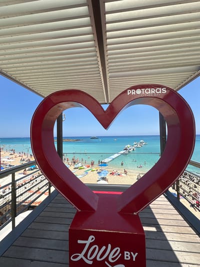 A vibrant red heart-shaped sculpture is positioned in the foreground, framing a picturesque view of Fig Tree Bay in Protaras, Cyprus. The sculpture, adorned with the words "PROTARAS" and "FIG TREE BAY" in white lettering at the top, and "Love BY" in cursive white lettering at the bottom, stands on a wooden deck. Beyond the heart, the turquoise waters of the bay teem with activity. Numerous people are visible on the sandy beach and swimming in the sea. A long, white jetty extends into the water, lined with several boats. The sky is a clear, brilliant blue, and the scene is bathed in bright sunlight, indicating a warm, sunny day. The overall impression is one of a popular tourist destination, offering relaxation and recreation.