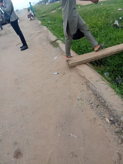 In Kagini, Nigeria, two individuals are captured on a dusty, uneven path adjacent to a strip of overgrown grass. To the left, a person in a light-colored jacket and dark pants walks away from the camera, carrying a black bag over their left shoulder. They are wearing dark shoes. To the right, another person, wearing a grey tunic and pants, is in mid-stride, stepping onto a weathered wooden plank that bridges a narrow drainage ditch. This individual wears dark sandals. The plank is positioned precariously over the ditch, which is filled with dark debris and some green vegetation. The ground is a mix of dry soil and small stones, with scattered pieces of white paper and other debris visible. In the background, more people, vehicles, and a building are faintly discernible, suggesting an urban or semi-urban environment.