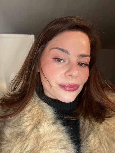 A woman with brown hair and light skin is angled with her head tilted to the right. She wears a black turtleneck sweater and a tan fur coat. Her makeup is natural with a glossy pink lipstick. Her eyes are brown, and her eyebrows are well-defined. The background is a plain, gray wall, and a portion of a white cabinet is visible in the lower-left corner.
