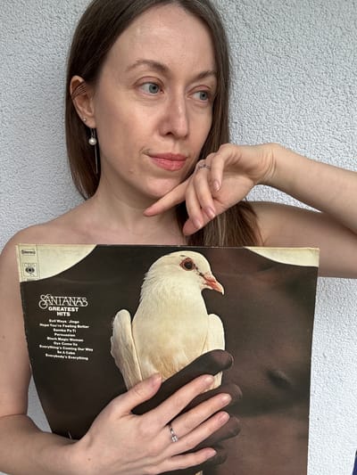 An adult woman is depicted lying down, holding a vinyl record sleeve against a textured white or light grey surface. She has long brown hair, fair skin, and light-colored eyes, looking upwards and to the side with a calm, contemplative expression. Her lips are slightly parted, enhanced with light pink lipstick. She wears a pearl drop earring in one ear and a small stud in the other, and rings are visible on her fingers. Her bare shoulder and arm are visible, suggesting a relaxed posture.

She holds a "Santana's Greatest Hits" vinyl record sleeve, with the album art showing dark-skinned hands cradling a white dove. Her right hand, adorned with a ring, rests over the dove on the record cover, while her left hand is positioned near her face. The album title "SANTANAS GREATEST HITS" and tracklist are clearly visible on the sleeve, including songs like "Evil Ways/Jingo," "Hope You're Feeling Better," "Samba Pa Ti," "Persuasion," "Black Magic Woman," "Oye Como Va," "Everything's Coming Our Way," "Se A Cabo," and "Everybody's Everything." The labels "Stereo," "CBS," and the catalog number "69081" are also discernible. The lighting is soft and natural, suggesting it is daytime. The general atmosphere is tranquil. The provided location context is Kudrovo, Russia.