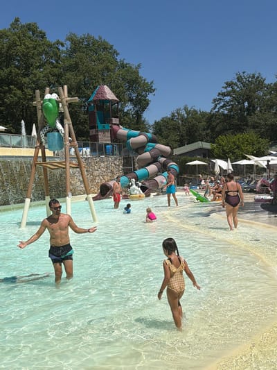 This image depicts a sunny day at a water park, likely within the Tuscan region of Italy, and specifically located in Cavriglia. In the foreground, a man stands in shallow, clear water, gesturing with his hands. Behind him, a young girl in a checkered swimsuit walks away from the camera, her hair in braids. Further back, several other individuals are visible, including a man in red swim trunks near a water slide, and a woman in a dark swimsuit walking away. The water park features several attractions, including a large, winding water slide structure with a playful design. A decorative element resembling a green frog with polka-dot legs is attached to a wooden structure. In the background, trees and umbrellas suggest a recreational outdoor setting. The water is shallow and sandy at the edges, with clear, illuminated water in the main area.