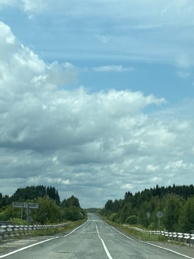 The image depicts a straight asphalt road extending into the distance, flanked by dense green trees on both sides. A white line divides the road, indicating two lanes of traffic. A metal guardrail runs along the right side of the road. In the distance, the road curves slightly upwards and disappears over a gentle hill. The sky above is a mix of blue with scattered white and gray clouds. A signpost with multiple signs is visible on the left side of the road, near the foreground. The scene appears to be a rural or semi-rural setting, likely on a highway or a main road, with no people or vehicles immediately visible.