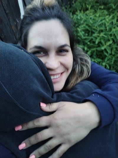 A young woman is captured in a close-up, seemingly hugging another person who is largely out of frame. She looks directly at the camera with a wide, joyful smile, revealing her teeth. Her dark hair, which appears to be pulled back, shows lighter, possibly highlighted, streaks around her face and shoulders. Her eyes are dark, complementing her olive skin tone. She is wearing a dark blue long-sleeved top or jacket.
Her hands are wrapped around the back of the person she is embracing, whose dark, possibly ribbed or corduroy-textured clothing is prominent in the foreground and center. The woman's fingernails are painted a vibrant coral pink, offering a striking color contrast against the dark fabric.
The background suggests an outdoor setting in Rusalevka, Russia. On the right, lush green foliage forms a soft, blurred backdrop, indicative of bushes or trees. On the left, a dark wooden structure, possibly a fence or part of a building, is partially visible. The lighting is soft and even, suggesting either an overcast day or the softer light of early morning or late afternoon. The overall mood conveyed by the woman's expression is one of happiness and affection. No text is visible in the image.