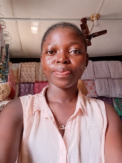 A single individual, a young adult female with dark skin, is depicted in a front-facing portrait, looking directly into the camera. She wears a light peach sleeveless collared shirt with visible buttons and a gold-colored necklace featuring a heart-shaped pendant. Her hair is pulled back, and small earrings are visible. The scene is an indoor environment, identified as a location within the city of Ayob, Nigeria. The background features various patterned textile fabrics, folded and stacked on shelves or racks. These fabrics display an array of colors including beige, brown, purple, pink, green, and white. To the upper left, red wire hangers are partially visible. The ceiling, appearing to consist of light-colored panels, features a brown, three-bladed ceiling fan with a white light bulb attached, positioned in the upper right portion of the frame. A portion of a decorated, possibly ceramic, object is visible on the far left, behind the subject's left shoulder. No other individuals are present in the frame.