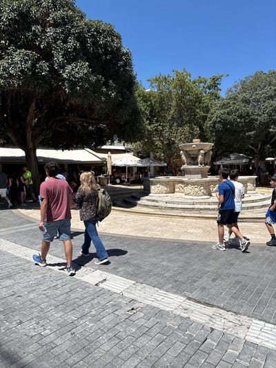 In Heraklion, Greece, a group of people walk on a cobblestone plaza in front of a decorative fountain. A large tree with dense foliage shades a portion of the scene. In the background, outdoor cafes with umbrellas and awnings suggest a lively public space. The sky is clear and blue. The people are dressed casually for warm weather. The fountain features intricate carvings and multiple tiers.
