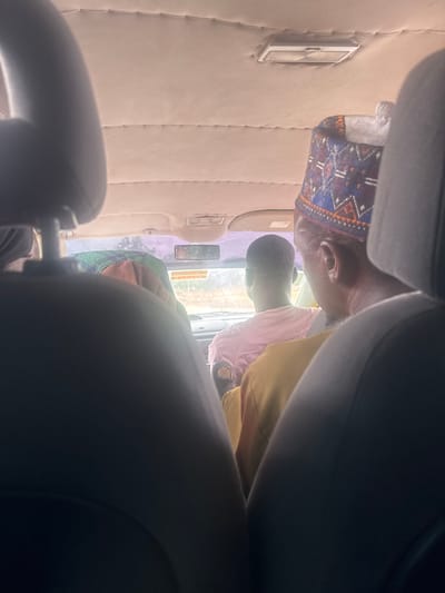 The image provides an interior view from the back seat of a vehicle, looking forward. The ceiling of the vehicle is light-colored and textured, with a rectangular light fixture visible. In the foreground, the backs of two dark car seats dominate the view.
Focusing on the occupants, there are at least three individuals visible. The person in the front right wears a patterned cap with blue, red, and white geometric designs. To their left, a person is wearing a pink shirt. Behind them, another individual is partially visible, wearing a yellow garment. The context of Kuru Dungus, Nigeria, is not directly discernible from the visual elements within the image itself. The lighting suggests it is daytime, and the view through the windshield indicates an outdoor environment, likely a road.