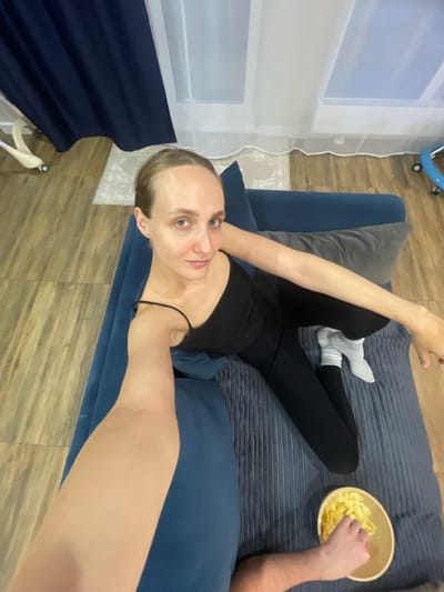 In a dimly lit room, a woman with blonde hair pulled back sits on a blue couch, wearing a black tank top and black leggings, with white socks. She is holding a phone in her left hand, taking a selfie. Her right arm is extended, and her hand is resting on the couch. A gray pillow is behind her, and to her right, a bowl of yellow snacks is on the couch. A hand is reaching into the bowl to grab a snack. The room has wooden floors and a blue curtain. The background shows a window with sheer white curtains. This scene is not specific to the city of Подшивалово, Russia, and provides no contextual clues for its location.