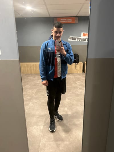 A single male subject is depicted in a full-body mirror selfie taken within what appears to be a changing room or common area of a sports facility in Hœnheim, France. The subject has short, dark hair and a light complexion, exhibiting a slight smile. He is dressed in a blue and dark blue zippered athletic jacket with a white Nike logo on the left chest, worn over a white, red, and black diagonally striped athletic shirt or jersey featuring "PSG" branding. Black athletic shorts are worn over black compression leggings, and he wears dark athletic shoes with white soles. His right hand holds a light-colored smartphone, positioned to capture the image, while his left hand is clenched into a fist.
The environment, visible in the mirror's reflection, consists of a grey wall with a light wood-paneled bench extending along its base and a light grey tiled floor. A black backpack rests on a section of the bench to the subject's right. An orange rectangular sign is affixed to the wall, displaying inverted white text, partially legible as "SERVIETTE OBLIGATOIRE" (Towel Required), indicating a facility rule. Overhead lighting is reflected in the mirror.