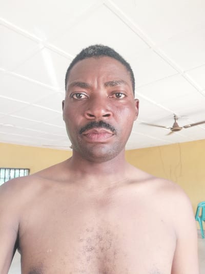 This media file is a close-up, front-facing portrait of a Black man. He is shirtless, with his shoulders and upper chest visible. The man has short, dark hair with some graying at the temples, dark eyes, and a mustache. His skin appears to be moist, suggesting he may be sweating.

The background is an indoor setting with a white, paneled ceiling. A ceiling fan is visible in the upper right portion of the frame. The walls appear to be a light yellow color, and there is a barred window or vent on the left side of the image. A turquoise chair is partially visible on the right side. The lighting suggests it could be daytime, possibly with bright natural light filtering in. There is no visible text in the image.

The man is looking directly at the camera with a neutral expression. The scene is static, with no obvious activity taking place beyond the static portrait. The location context provided is Ajasse Ipo, Nigeria.