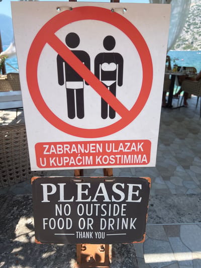 The image shows two signs posted outdoors, likely in Risan, Montenegro, given the context of coastal areas and multilingual signage. The upper sign features a red prohibition circle with a diagonal line over two black silhouette figures. One figure appears to be male, and the other, which is to the right, is depicted wearing a bikini top and bottom, indicating a restriction on wearing swimwear. Below this graphic, in a red rectangle, is text in a language other than English, which reads "ZABRANJEN ULAZAK U KUPAĆIM KOSTIMIMA". This translates to "Entrance forbidden in swimsuits". The lower sign is rectangular with a dark brown background and white text. It reads "PLEASE NO OUTSIDE FOOD OR DRINK THANK YOU". The signs are mounted on a metal post, and the background shows a blurred view of a coastal area with tables and chairs, suggesting a restaurant or cafe by the sea. The ground is paved with stone tiles, and there are some rocks and shadows present, indicating it is daytime. There are no visible people interacting directly with the signs.