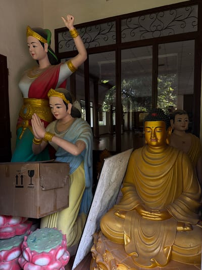 The photograph, taken in Thủy Xuân Ward, Vietnam, shows several Buddhist statues. The largest is a golden Buddha in a seated meditative pose, placed near a window with decorative ironwork. Behind the Buddha is a smaller figure with a swastika symbol on its chest. In the foreground, two statues depicting female figures are positioned side-by-side. One figure extends her arm upwards, while the other clasps her hands together in a gesture of prayer. A cardboard box labeled "FRAGILE" partially obscures the second female figure and a decorative lotus base.