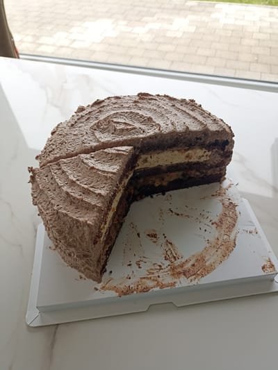 A chocolate cake with a slice removed sits on a white cardboard base, which is resting on a marble-patterned countertop. The cake is covered in textured chocolate frosting, resembling tree bark. The cut reveals multiple layers of dark chocolate cake interspersed with lighter colored cream and nut-filled fillings. Crumbs and smears of chocolate frosting are visible on the cardboard base and the countertop. In the background, through a window, a paved outdoor area with some greenery is visible. The lighting suggests an indoor setting, likely a home or a cafe. The image does not contain any information or context related to the location in Doirí Beaga, Ireland.