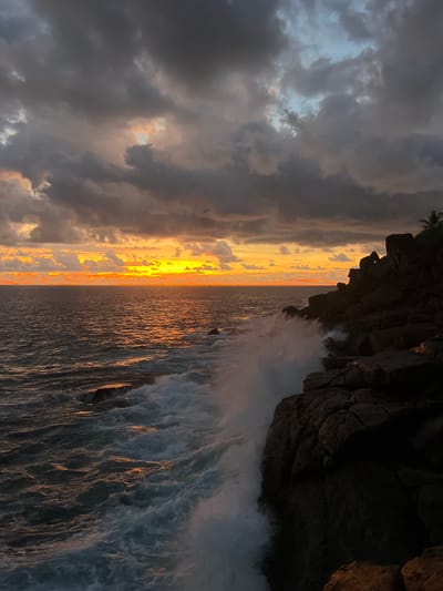 This photograph captures a dramatic coastal landscape at either sunrise or sunset, likely in Rumassala, Sri Lanka. The scene is dominated by a clear horizon line where the sky meets the ocean.

On the left, the sky is filled with dark, heavy clouds that create a moody and intense atmosphere. However, a brilliant golden and orange light, presumably from the sun, breaks through a patch in these clouds, casting a fiery glow towards the horizon. Hints of lighter, possibly blue, sky are visible through thinner cloud cover.

The center of the image is defined by the ocean, which intensely reflects the golden light, creating a shimmering path of bright orange and yellow hues extending towards the horizon. The water surface shows visible textures of waves and currents, some catching the warm light.

To the right, dark, rugged rocky cliffs rise prominently, casting a striking silhouette against the illuminated sky and ocean. At the very top of these cliffs, a small portion of a palm tree or similar tropical vegetation is visible. Large, powerful waves are actively crashing against the base of these cliffs, generating significant white foam and sea spray that adds to the dynamic nature of the scene.

The overall impression is one of natural power and dramatic beauty during the golden hour, with the contrasting dark clouds and bright sunlight creating a captivating visual experience. No visible text is present in the image.