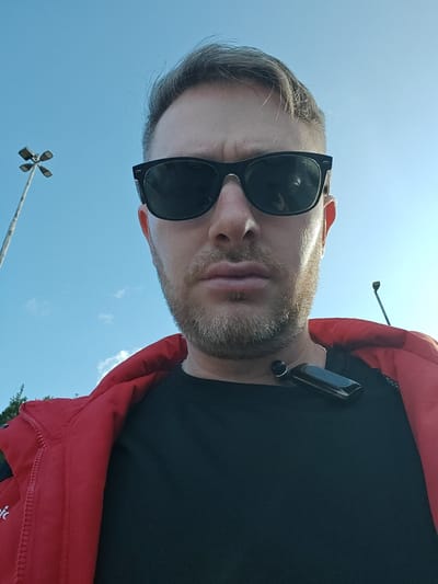 A male individual is depicted in a close-up, low-angle selfie, outdoors in Nesher, Israel. The subject has light-colored hair and a beard, and is wearing black sunglasses, a black t-shirt, and a red jacket. A black, rectangular wearable device with a round component is situated on his neck, partially obscured by his right ear. His lips are slightly pursed. The background reveals a clear blue sky with minimal, wispy white clouds. Two streetlights are partially visible: one multi-headed fixture on the far left, and a single-headed fixture on the far right. Slight green foliage is present in the lower-left corner.