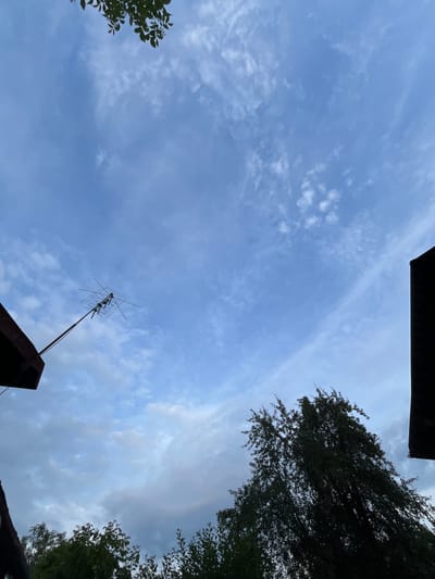 The image depicts a sky with scattered clouds, a television antenna, and the tops of trees. Two dark, angled structures, possibly rooftops or eaves, are visible on the left and right edges of the frame. The scene is shot from a low angle, looking upwards. There are no people or visible actions taking place. The overall impression is that of an outdoor setting, possibly a residential area.