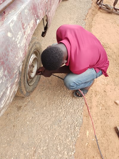 In Serti, Nigeria, a person wearing a crimson shirt and blue jeans is seen crouching next to the rear tire of a weathered, maroon vehicle. The individual is using an air hose to inflate the tire. The ground is a mixture of gravel and dirt, with the vehicle's tire resting on the gravel. The vehicle's body appears to be in disrepair, with visible rust and peeling paint.