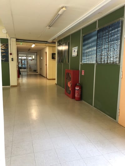 The image displays an empty, well-lit hallway or corridor, likely within a school or institutional building in Agia Paraskevi, Greece. The perspective looks down the length of the corridor, which features a light-colored, tiled floor that reflects overhead lighting and natural light from further down.
On the right side, the wall is comprised of green panels, possibly blackboards or decorative elements, framed by lighter strips. Integrated into this wall are several transparent or reflective panels, showing distorted reflections of light and patterns. A poster affixed to one of these green panels partially reads "EUROPEAN SCHOOL SPORT DAY." Below this, a red fire hose cabinet is mounted, and a red fire extinguisher stands upright on top of it. Text visible on the fire extinguisher includes "PRESTO" and "KG," along with some Greek characters. A white sign with a red arrow pointing right is positioned above the fire hose cabinet.
The left wall of the hallway is a plain, light-colored surface, featuring a long, white fluorescent light fixture mounted horizontally. Further down the corridor, the space appears to lead towards glass doors or partitions, through which additional light enters the area. No individuals are present, suggesting the hallway is quiet, perhaps during off-hours or a break. The overall impression is one of a functional, clean, and organized institutional environment, typical of a school building.