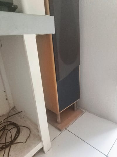 An indoor scene in Cimenyan, Indonesia, presents a static arrangement of objects within a room. On the left side of the frame, a portion of a white, presumably wooden, shelving unit or desk is visible, featuring a top and a lower shelf. On the lower shelf, a dark-colored electrical cable is loosely coiled. Adjacent to this unit, positioned towards the right against a light-colored wall, stands a floor-standing audio speaker. The speaker cabinet is light brown or wood-toned, with the upper and lower sections of its front face covered by dark grey or black grille cloth. It rests on a separate, lighter wooden base supported by four short, cylindrical feet. The floor is composed of light-colored, possibly white, ceramic tiles with visible dark grout lines. No individuals are present, and no active interactions are depicted. The area on the lower shelf, beneath the coiled cable, appears to have accumulated dust or grime.