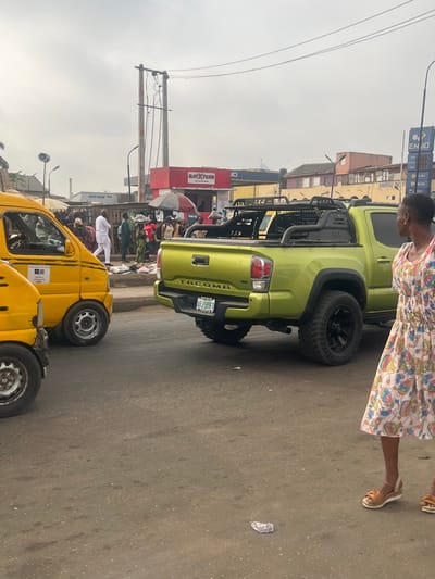 The image portrays a bustling urban street scene in Egan, Nigeria, under an overcast sky. In the foreground to the right, a person with dark skin and short hair, dressed in a light-colored patterned dress and brown sandals, stands facing left, observing the activity. A lime green Toyota Tacoma pickup truck occupies the central mid-ground, featuring large black off-road tires and a black roll bar/bed rack system. Its Nigerian license plate, ABJ 599FL, is affixed to the rear. To the left of the pickup, two yellow, compact public transport vans are partially visible, one behind the other. The asphalt road surface appears worn, with minor debris present.
The background reveals a lively commercial area. Numerous pedestrians are visible on the sidewalk, including a male in a white traditional garment walking leftward and another individual carrying items on their head. Commercial establishments include a red building with "SLOT XPRESS" signage and an associated umbrella-shaded stall, and a prominent blue structure with a tall sign displaying "ENJO" and pricing information, indicative of a petrol station. Utility poles with multiple wires and streetlights are interspersed throughout the scene. Buildings of various architectural styles complete the urban backdrop.
