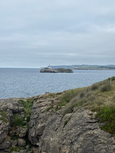 This is a photograph of a lighthouse on a rocky outcrop in the sea. The lighthouse is white and cylindrical with a lantern room at the top. The rocky outcrop is dark and irregular in shape, and it is surrounded by blue water. In the foreground, there is a rocky cliff with green vegetation. In the distance, there is a coastline with rolling hills. The sky is overcast and gray. This scene is likely located at Cabo Mayor in Santander, Spain, a prominent cape known for its lighthouse and dramatic coastal cliffs. No people are visible in the image.