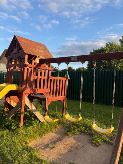 In Мещёры, Russia, a wooden children's playground set stands outdoors on a grassy area. The structure is red and features a red-tiled roof, a yellow slide, and a wooden ladder. Three yellow swings hang from a horizontal wooden beam. A green metal fence runs along the background. The sky is blue with scattered white clouds, and the sun is casting long shadows.