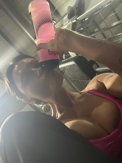 This image depicts an adult female mid-action, drinking from a pink Science in Sport (SiS) branded water bottle, held in her right hand. Her head is tilted upwards, and she wears a fuchsia athletic top and dark lower garments. A visible sheen indicates moisture on her skin. The background reveals industrial overhead structures, including parallel silver metallic bars and dark utility fixtures, alongside a partially visible black padded section of gym equipment positioned behind her right shoulder. The environment is an indoor fitness facility located in Tunbridge Wells, United Kingdom.