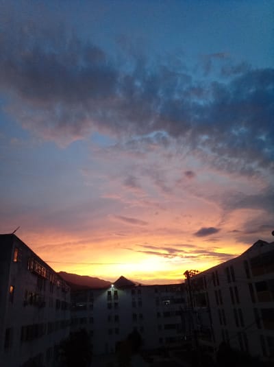 The image depicts a sunset in Parroquia Guatire, Venezuela, with buildings framing the view of the sky. The sky displays a gradient of colors, from deep blue at the top to orange and yellow near the horizon. Clouds are scattered throughout the sky, with darker, gray clouds in the upper portion and lighter, pink-tinted clouds closer to the horizon. Below the sky, the silhouettes of buildings are visible on either side of the frame. The buildings are dark in tone, and their architecture is visible. In the distance, a dark mountain range is just above the buildings. There is no evidence of people, subjects, or actions in the image.