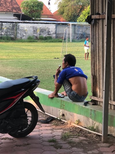 An adult male is sitting on a green and concrete wall, smoking a cigarette. He is wearing a blue t-shirt, pulled up to expose his back, and denim jeans. His left leg is bent, and his right leg is tucked underneath him. He is holding a phone in his right hand. In the foreground, the rear of a black and red motorcycle is visible. To the left of the wall, a pair of black flip-flops are on the ground. In the background, a grassy field with a soccer goal can be seen, along with a young boy in a white t-shirt and yellow shorts standing near the goal. Trees and buildings are visible behind the field. The scene appears to be an outdoor area, possibly a sports field or park, in Pamotan, Indonesia.