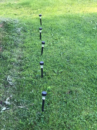 The image shows a row of six solar-powered garden lights planted in a grassy area. The lights are black and silver with clear tops. The grass is green and appears to be recently mowed in some areas, with some clippings visible on the left side of the frame. The scene is outdoors, with natural lighting suggesting it is daytime. The specific location in the city of Zhukovo, Russia, cannot be determined from the image alone. There are no people or animals visible in the photograph.