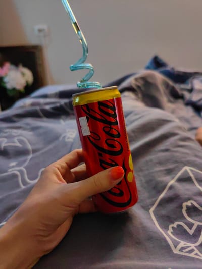 A person's hand, with bright orange nail polish, holds a red and yellow can of Coca-Cola. A blue, coiled plastic straw extends from the top of the can, bent in a spiral shape. The can has the Coca-Cola logo prominently displayed in black. A small white sticker with black text, possibly a price tag, is adhered to the can. The background consists of a bed with gray bedding that has a white outline pattern of cartoonish figures. The scene appears to be indoors, likely in a bedroom. There is no discernible information within the image to indicate the location in the city of Fanipal, Belarus.