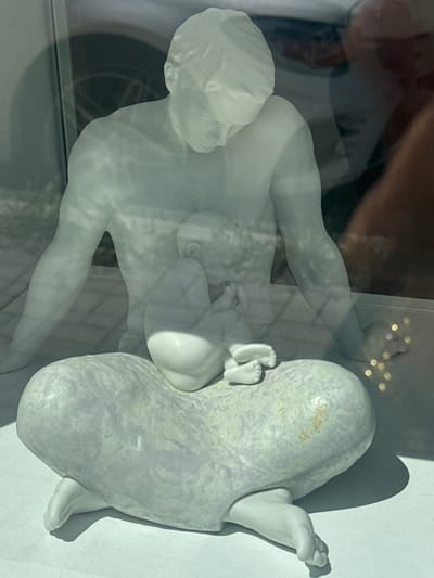 This is a close-up, vertically oriented photograph of a white ceramic sculpture. The sculpture appears to depict a human figure, possibly a mother and child. The main figure is lying down, with their legs extended. A smaller figure, presumably a baby, is nestled against the larger figure. There are hints of a reflection in the surface the sculpture is resting on, suggesting it might be displayed behind glass. The lighting is bright, casting shadows that add depth to the sculpture. The background is white and appears to be a studio or exhibition setting. The sculpture has a smooth, matte finish with some subtle variations in tone. The overall impression is one of tenderness and maternal care. There is no visible text.