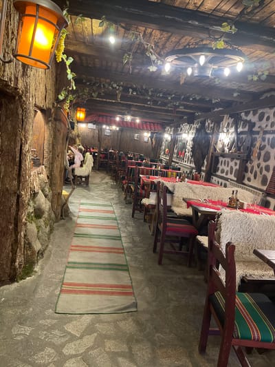 The image captures the interior of a restaurant in Bansko, Bulgaria. The space is adorned with rustic wooden structures, including a wall made of what appears to be stacked logs and a ceiling with a pattern of stones. There are vines and decorative lights along the wooden structures, creating a warm ambiance. Tables with fur-like covers are lined along a corridor. One table is set with glassware and bottles. Chairs with striped cushions surround the tables. A rug with vertical red and green stripes runs along the corridor. In the distance, there are people seated at a table. A prominent feature is an illuminated lantern in the foreground, casting a bright orange glow.