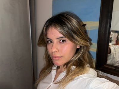 A woman with brown hair and highlights is positioned in a room. She wears a white collared shirt. A painting hangs behind her, its colors predominantly blue and beige. A dark-framed mirror is also visible to the right. A refrigerator is placed on the left side of the photograph. The woman appears to be the primary subject, and the setting includes interior elements suggestive of a domestic space. The location, Sumalao, Argentina, is not visibly evident from the image itself.