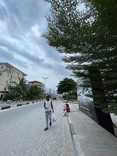 In Ilado, Nigeria, a man wearing a white sleeveless vest and grey pants walks on a paved road carrying a white plastic bag in his right hand and a phone in his left. He is heading towards the viewer, slightly to the right. Behind him, to the left, are several buildings and palm trees. To the right, a large black billboard with text and an image stands beside a sidewalk where two people are sitting. The sky is cloudy.