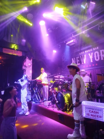 In Patong, Thailand, a live music bar called New York is hosting a performance. The stage is illuminated by purple and yellow lights, creating a vibrant atmosphere. A female singer, dressed in white, holds a microphone and sings to the audience. Beside her, a male keyboardist, also in white, plays a red keyboard. In the background, a drummer is seated behind a drum set. A male performer in a white hat and sleeveless shirt, holding a microphone, stands in the foreground, facing the audience. A sign in front of him reads "CLAPPING IS NICE CASH IS NICER." Two women are visible in the lower left of the image, one singing and the other watching the performance. The scene suggests a lively and energetic evening at a music venue.