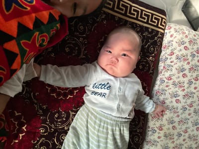 In this upside-down image, two individuals are lying on a patterned surface. The primary subject is a baby with a round face and dark eyes, wearing a light-colored onesie with green and white stripes on the upper part. The onesie has blue text on the chest that reads "little BEAR". The baby's hands are visible, one resting on a floral patterned fabric. To the right of the baby, another person's face is partially visible, also lying down. The background consists of a dark maroon fabric with an intricate red and gold floral pattern, and a section of a colorful, patterned blanket or garment. The setting appears to be indoors, possibly a bed or floor, with the overall impression being one of relaxation or rest. The lighting suggests it could be daytime.