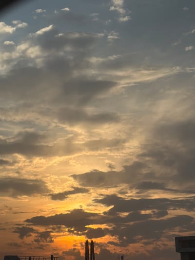 The image depicts a sky filled with clouds, with hues transitioning from blue to orange, suggestive of either sunrise or sunset. The scene includes structures, possibly buildings, visible on the right side of the image. The location is potentially Ferno, Italy, as specified in the request.