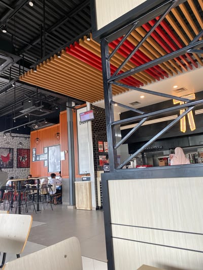 The image depicts the interior of a KFC restaurant in Lhoksukon, Indonesia. The restaurant features a modern design with a black metal frame structure in the foreground, partially obscuring the view of the seating area. The ceiling has a slatted wooden and red design. In the background, customers are seated at tables. A person wearing a light pink hijab is visible behind the counter, indicating the presence of staff. The walls are decorated with black and white patterned wallpaper and framed artwork. A digital menu board is also visible. The overall environment suggests a well-lit and operational fast-food establishment.