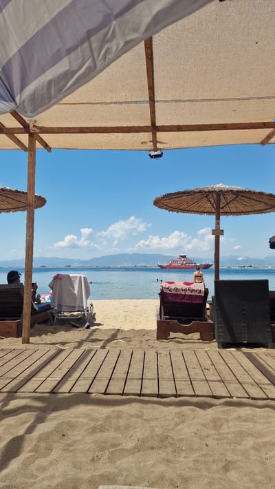 The image depicts a sunny day at the beach in Keramoti, Greece. In the foreground, a wooden boardwalk leads towards the sandy beach. Several sun loungers, some occupied by individuals, are visible, offering shade under thatched umbrellas. In the background, the tranquil sea stretches to the horizon, where a large red and white ferry boat is seen sailing. Distant hills and a clear blue sky with scattered white clouds complete the scenic view.